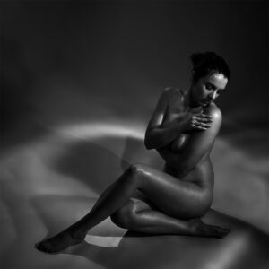 Fine art nude photograph of a seated female figure looking downward, illuminated by soft light and shadow