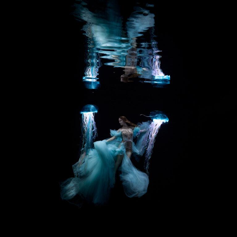 Woman in turquoise haute couture gown floating underwater with two glowing LumiJelly props, real water surface reflections visible above – single shot, no composite, no CGI. Underwater fashion photography by Stephan Ernst, gown by Oksana Schleekeil
